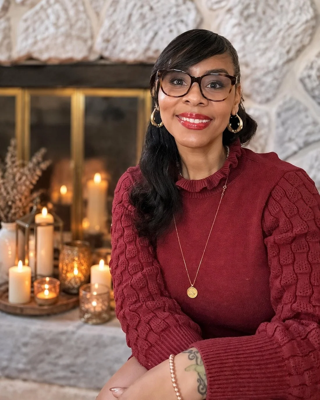 Misha Williams in glasses and a burgundy sweater, smiling in front of a fireplace lit with candles