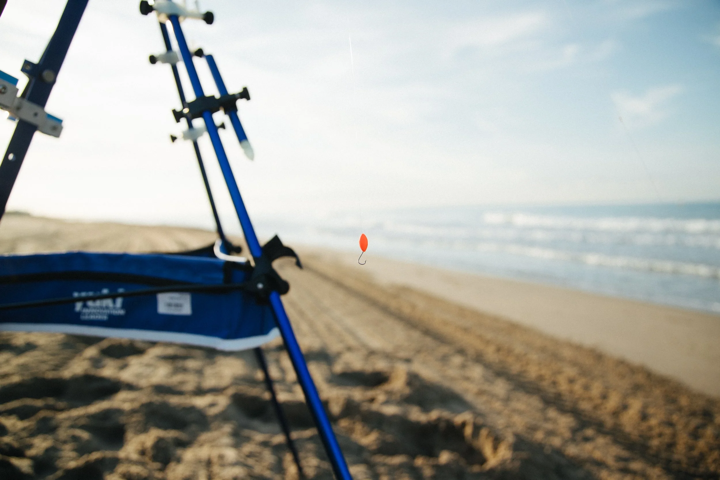 A beach with a fishing pole and an orange fishing bobber floating in the air, with the ocean and sky in the background.