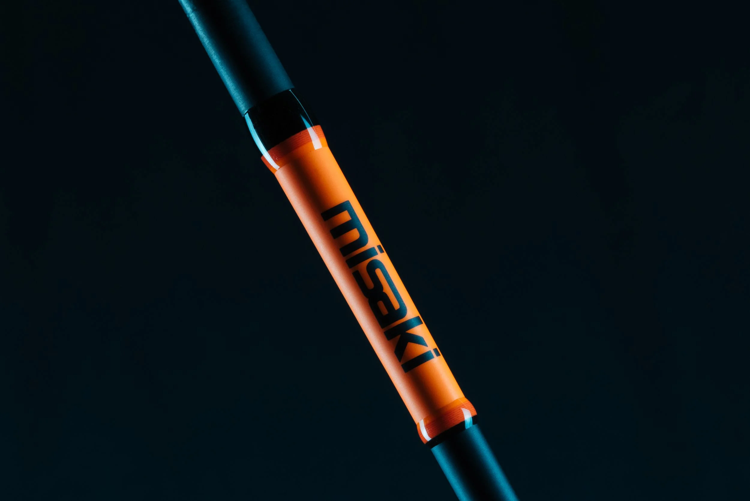 Close-up of a black and orange tennis racket handle with the brand name "MISHKI" in bold letters, set against a dark background.