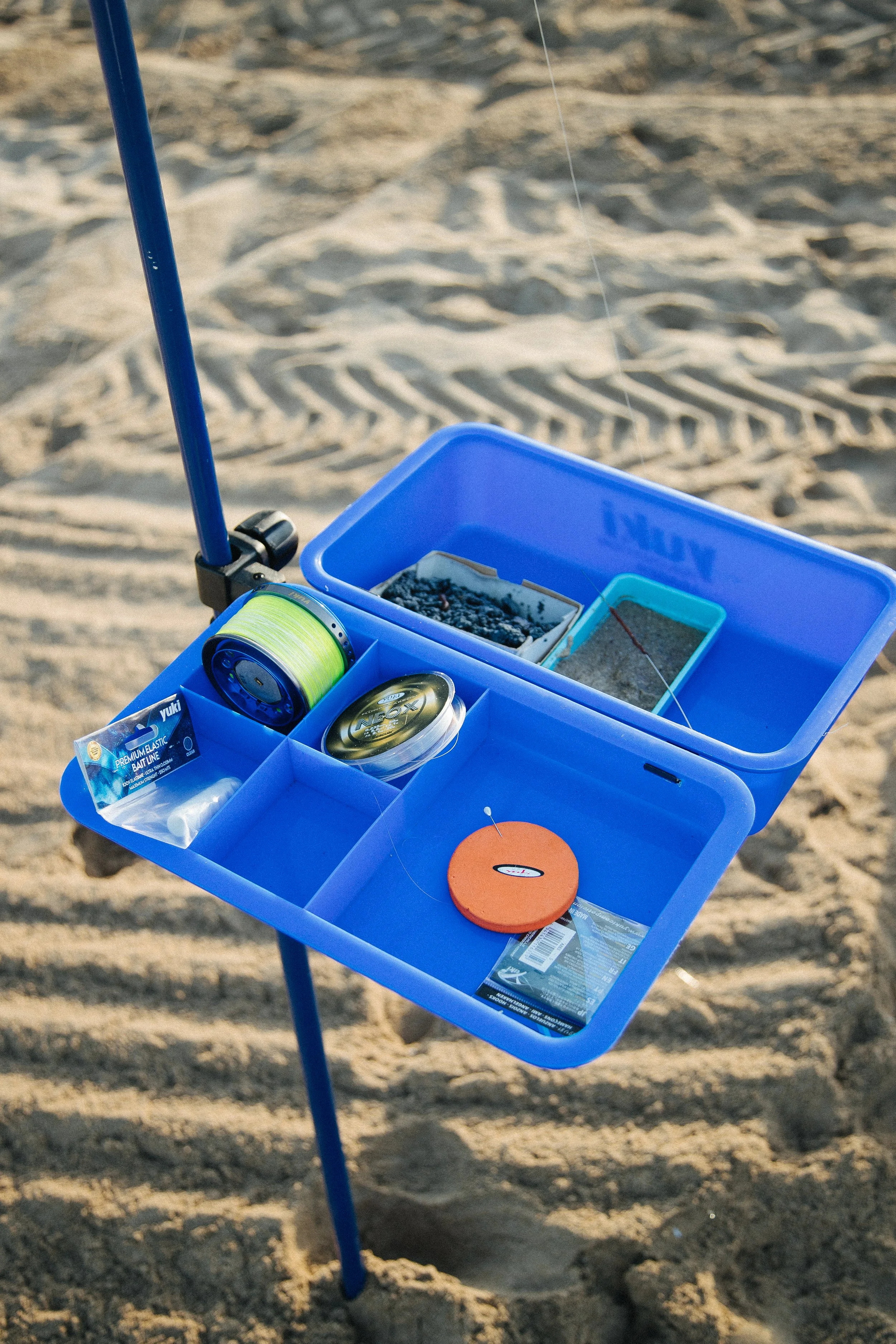 Blue tackle box on sandy beach with fishing gear and bait.