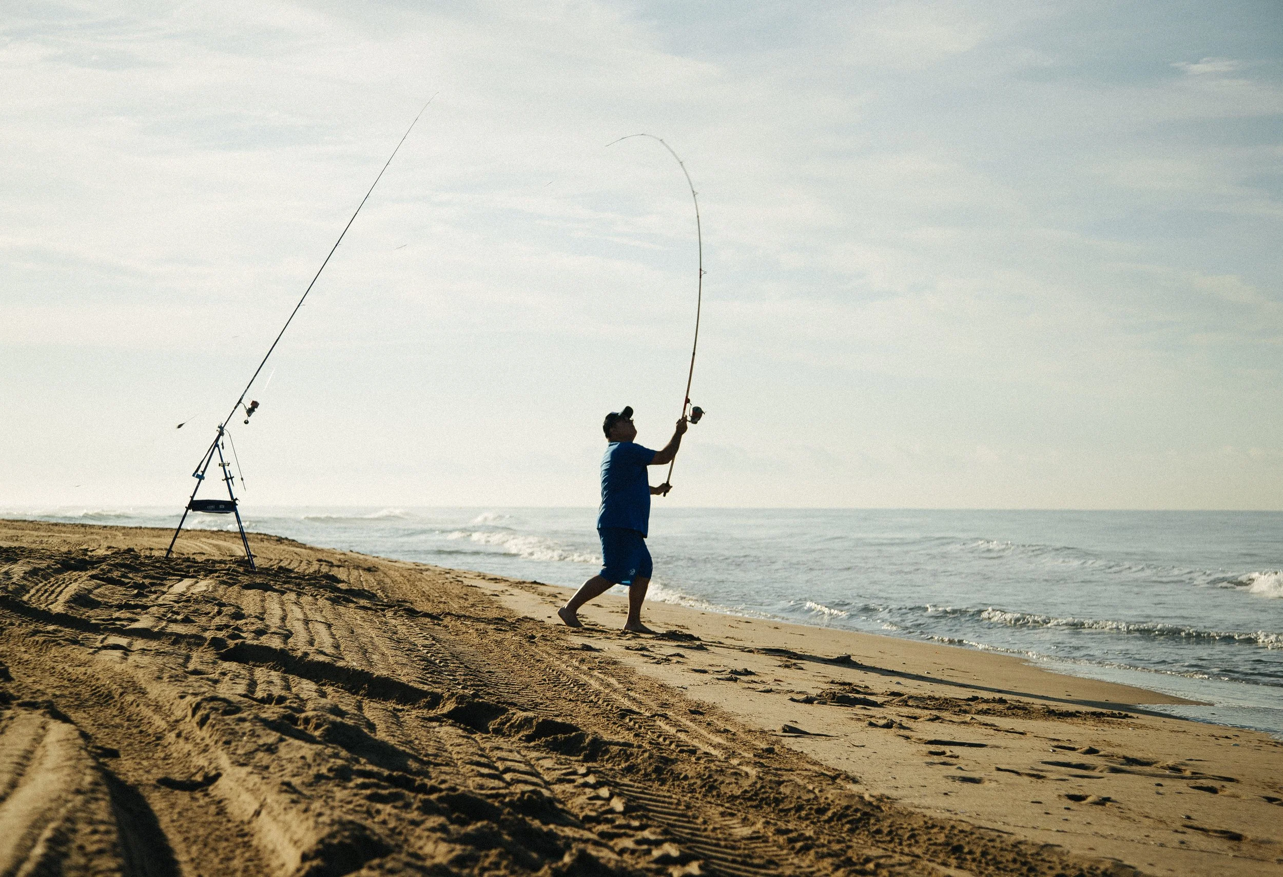 Person in blue shirt and shorts fishing on a sandy beach near the ocean, with multiple fishing rods set up behind them.