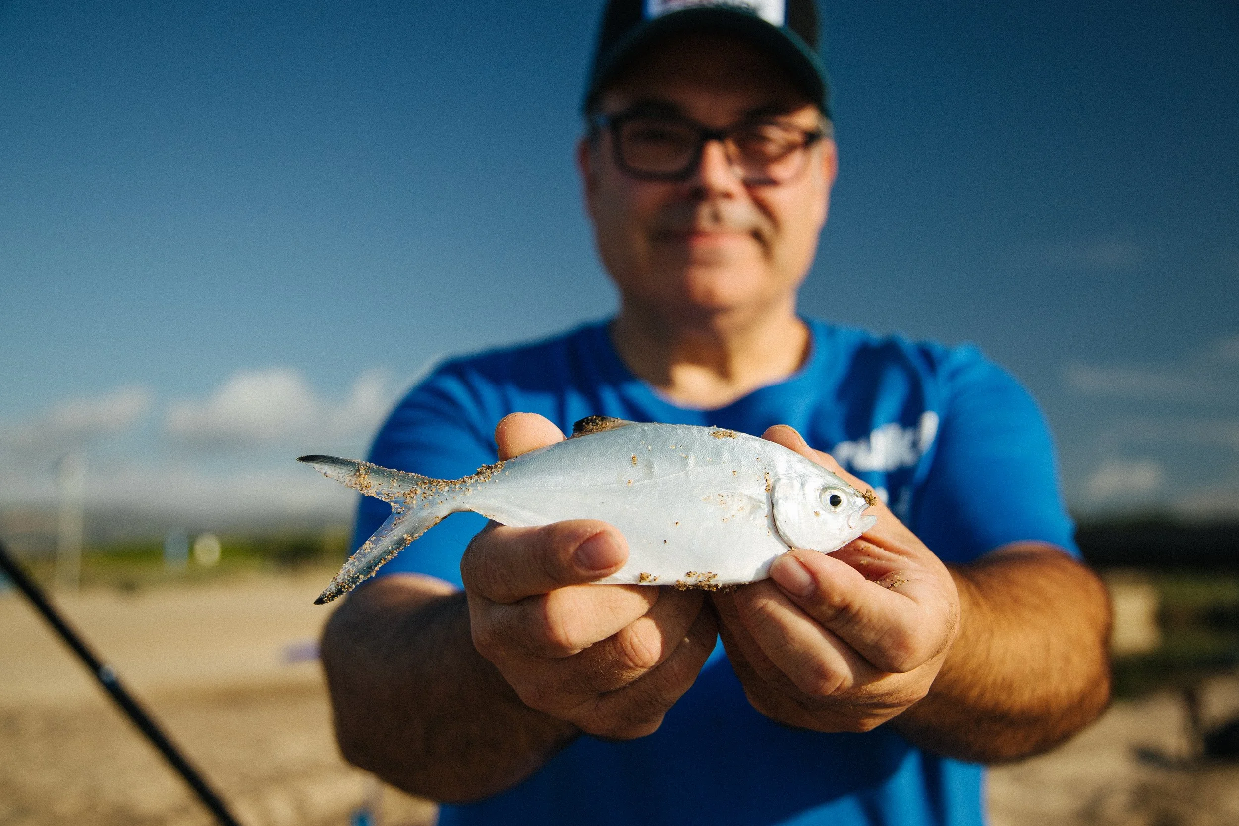 Man holding a small fish on a beach, wearing glasses and a blue shirt, with a fishing rod visible in the background.