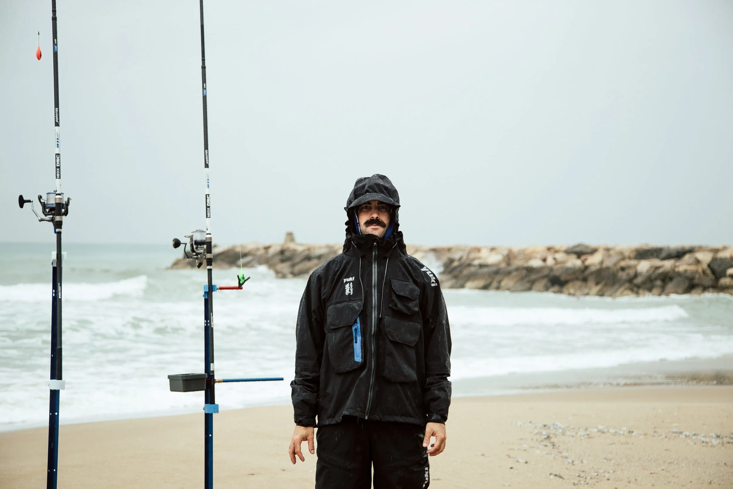 Man in a black waterproof jacket and hat standing on a beach, fishing poles set up next to him, with rocks and ocean waves in the background.