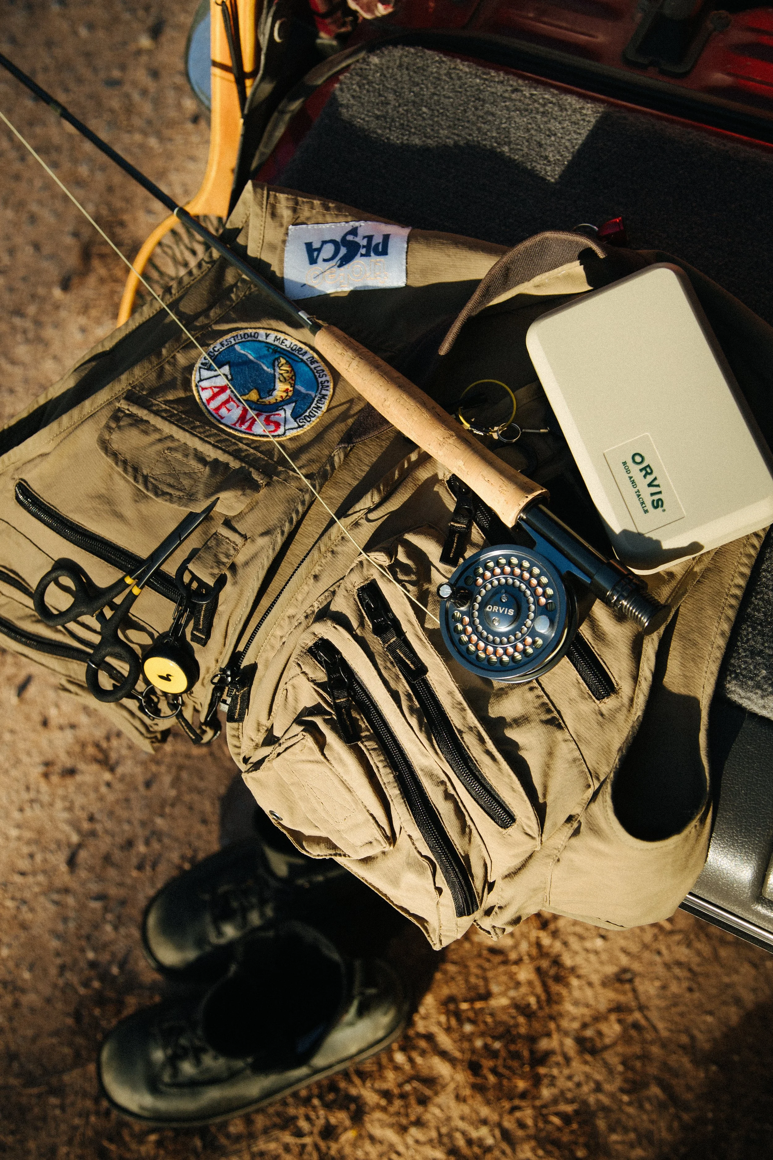A fishing gear setup in a bag on a vehicle with a pair of boots on the ground nearby, including a fishing rod, a fishing reel, scissors, and other tackle.