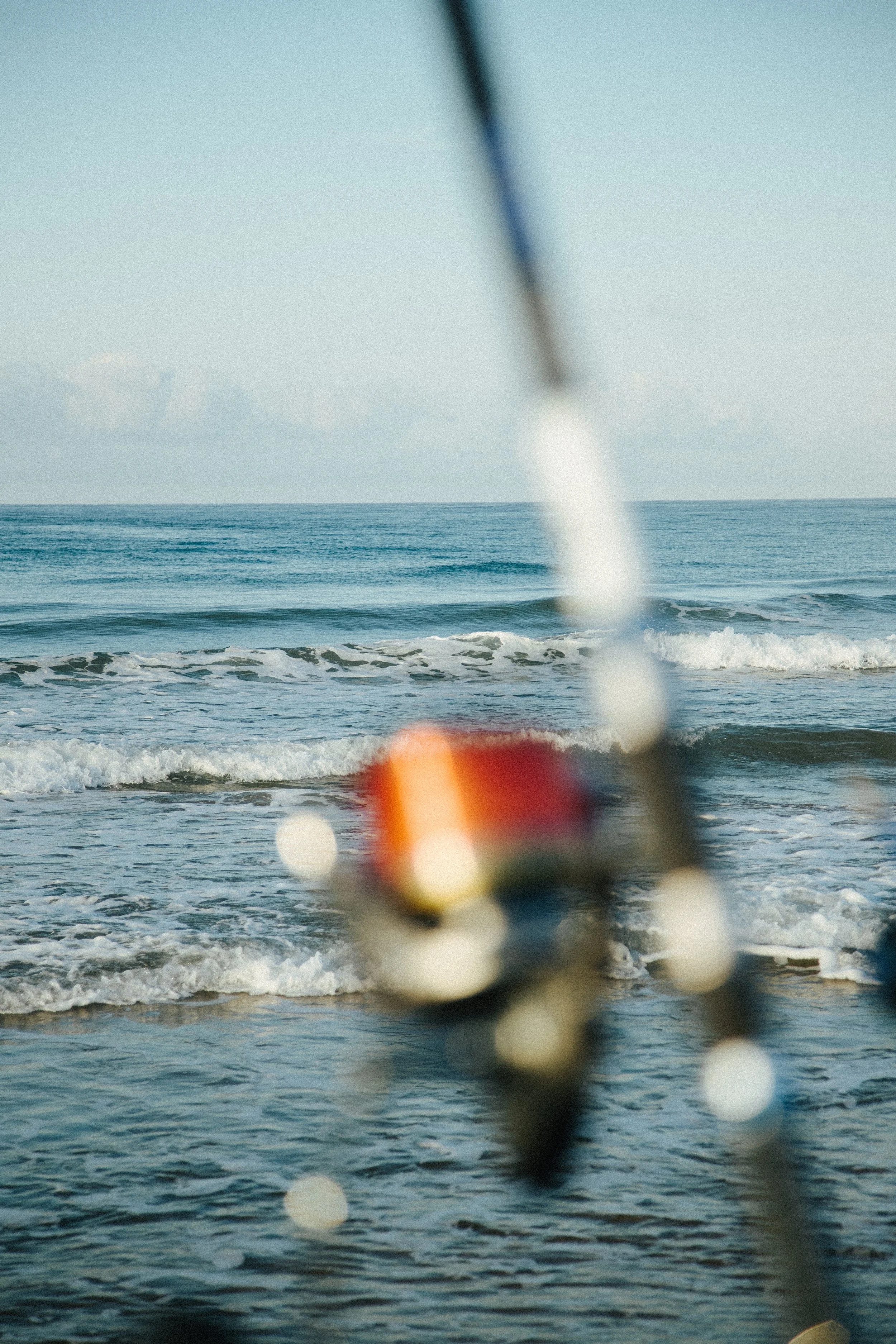 Blurred fishing rod with colorful reel in front of a calm ocean with gentle waves and a clear sky.