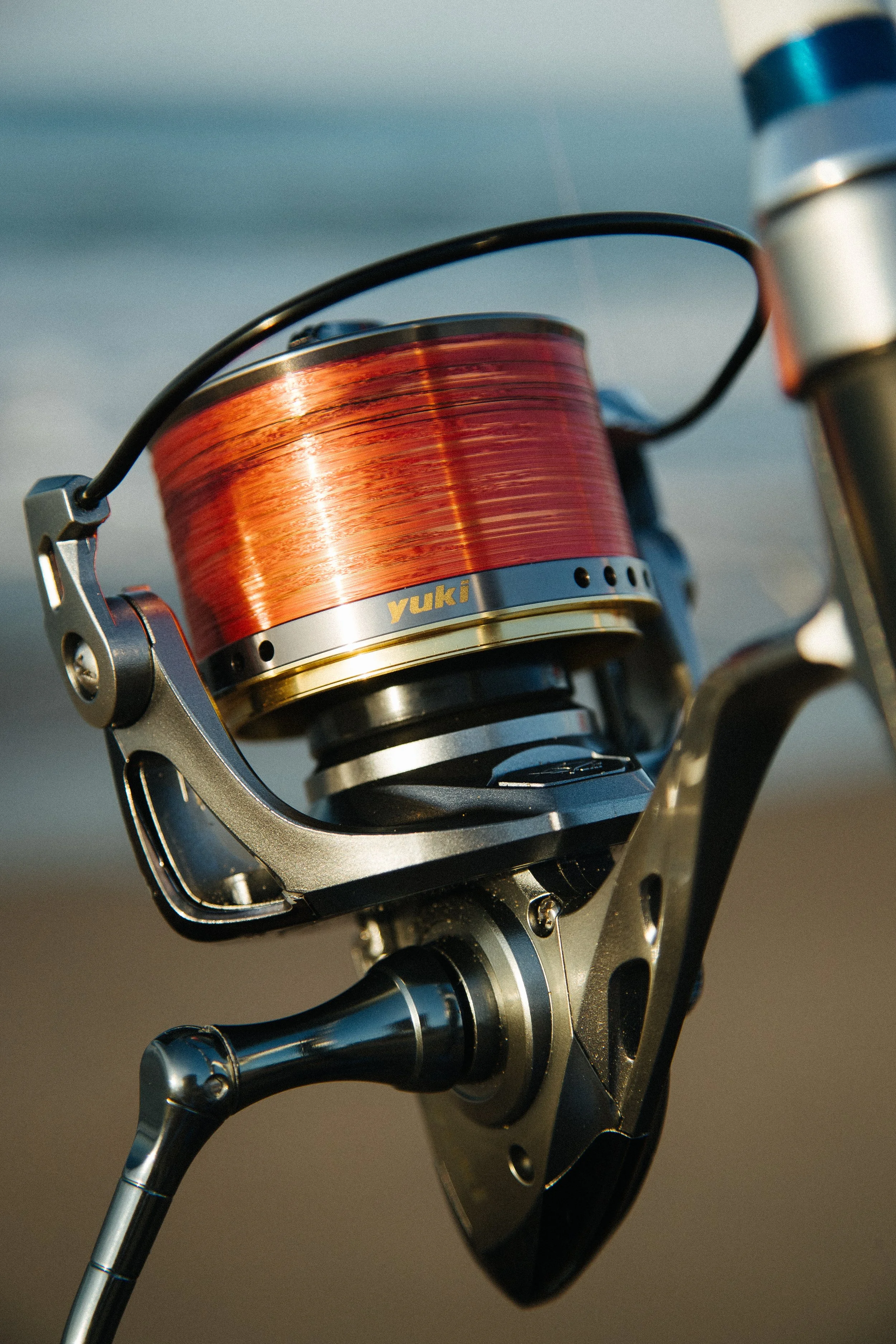 Close-up of a fishing reel with orange fishing line, attached to a fishing rod, on a beach or near water.