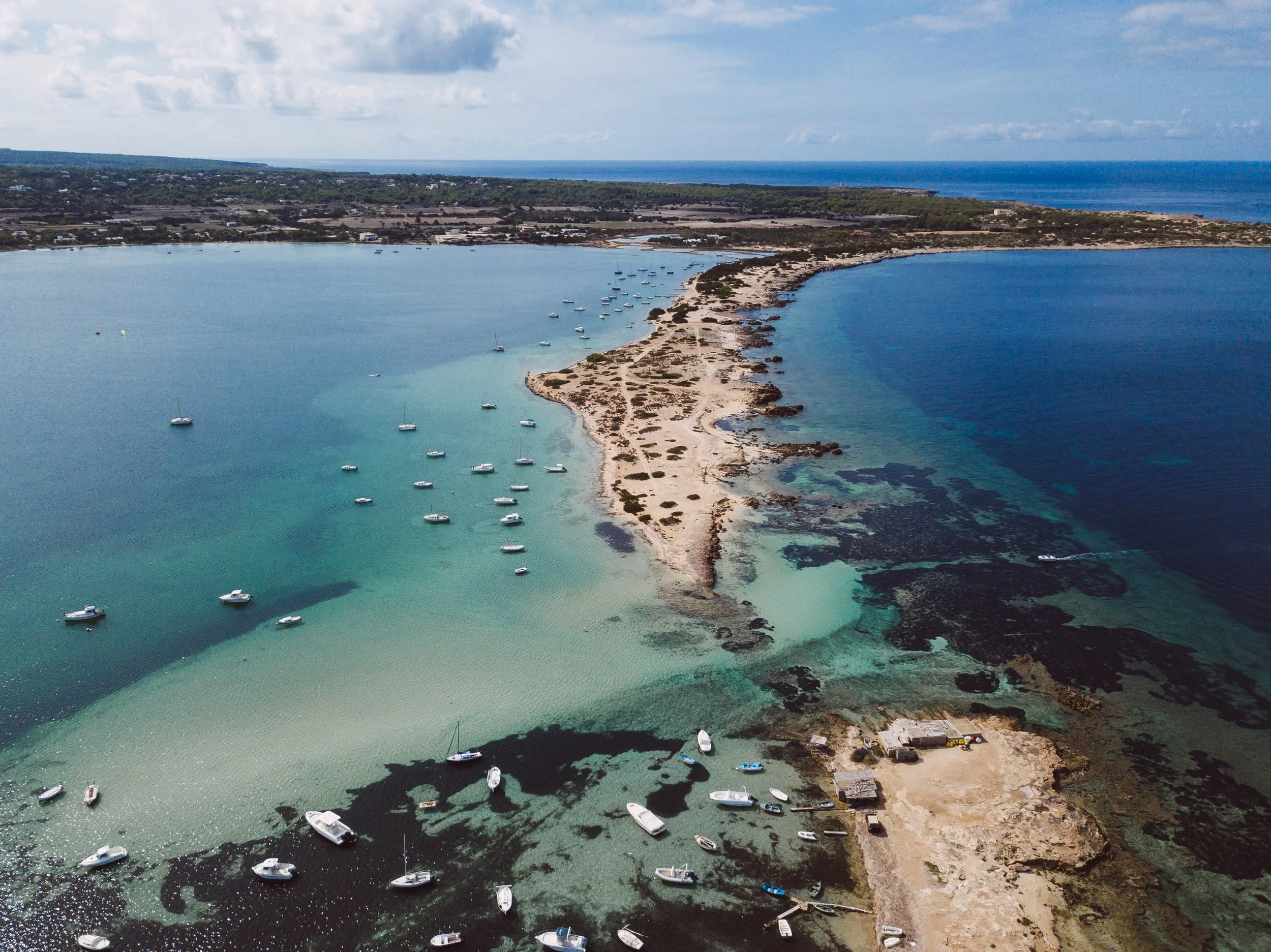 Aerial view of a narrow sandbar connecting two land masses, with boats anchored in the surrounding turquoise and dark blue waters, and a distant shoreline with scattered buildings and greenery.