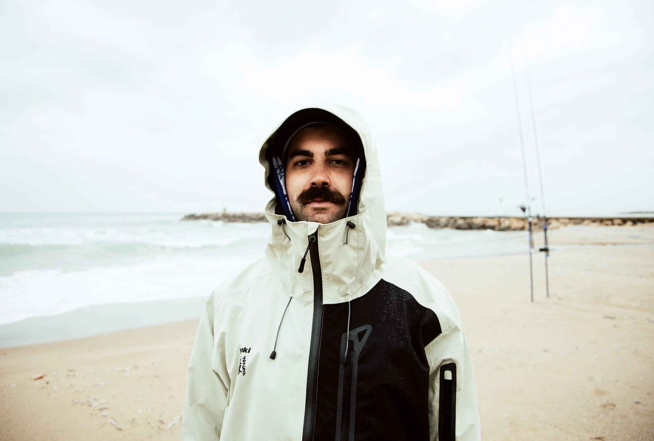 Man in a beige and black waterproof jacket standing on a beach with fishing poles in the background on a cloudy day.