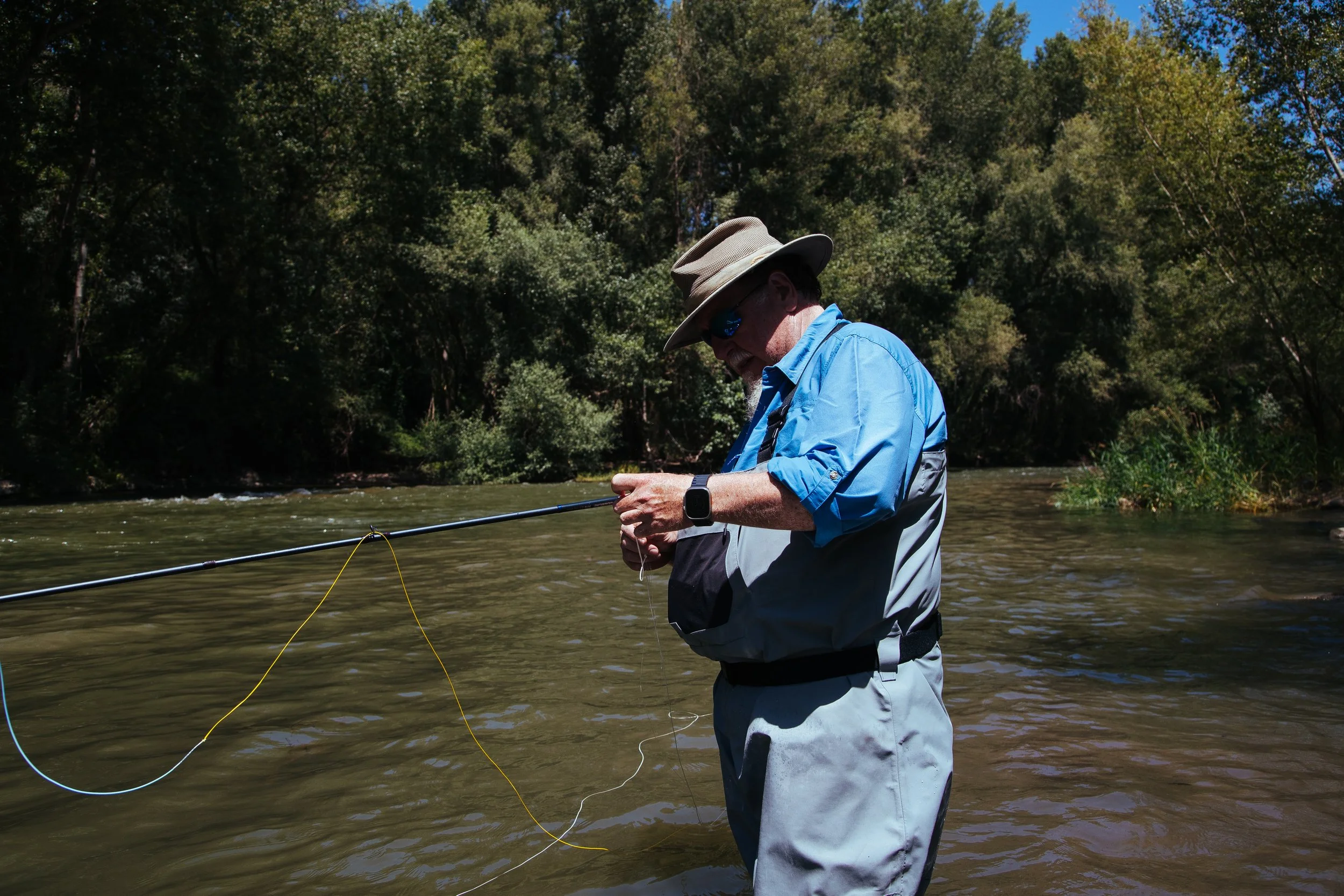 Older man standing in a river, fishing with a rod, surrounded by trees.
