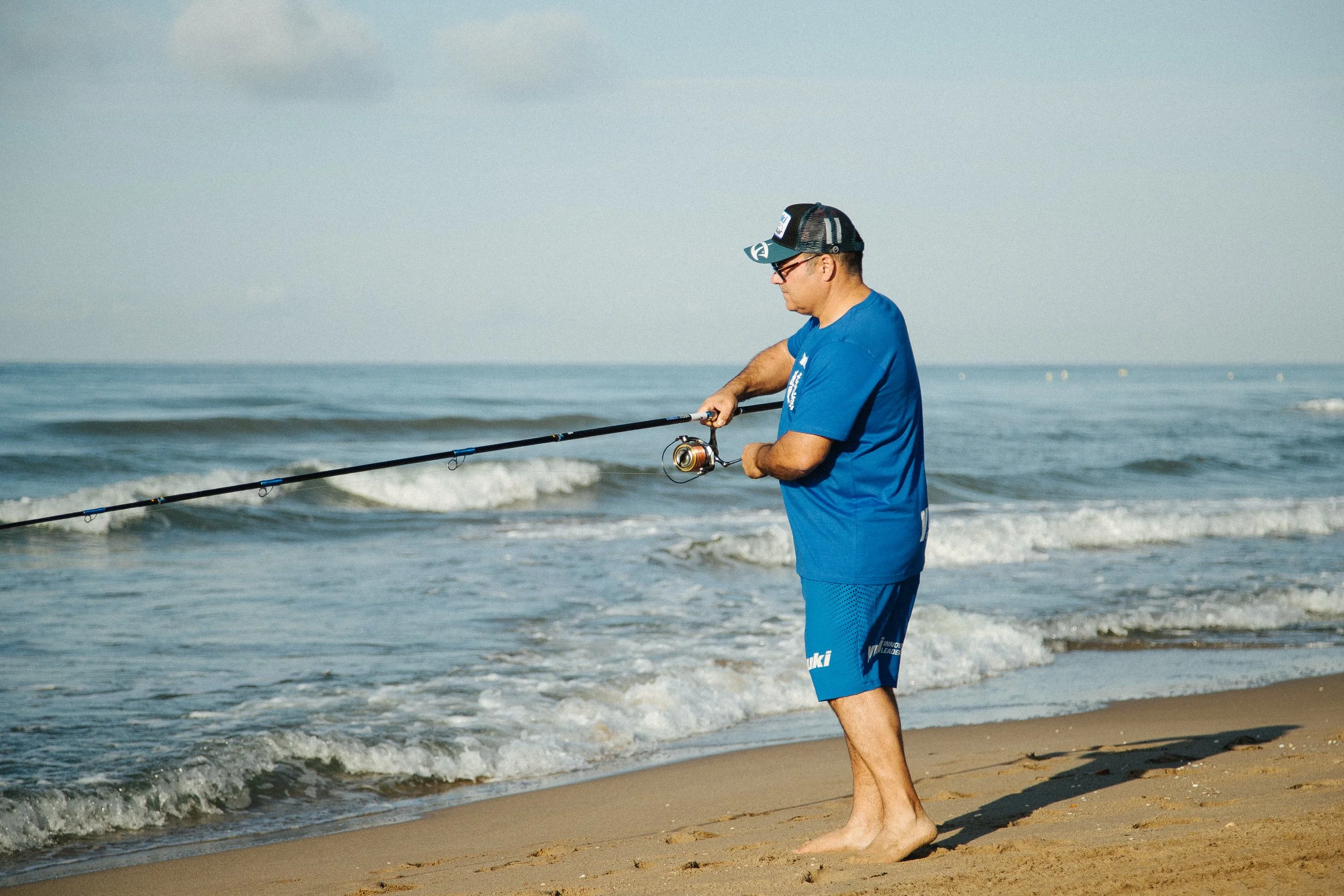 A man in a blue t-shirt and shorts fishing on the sandy beach by the ocean, with small waves in the background.