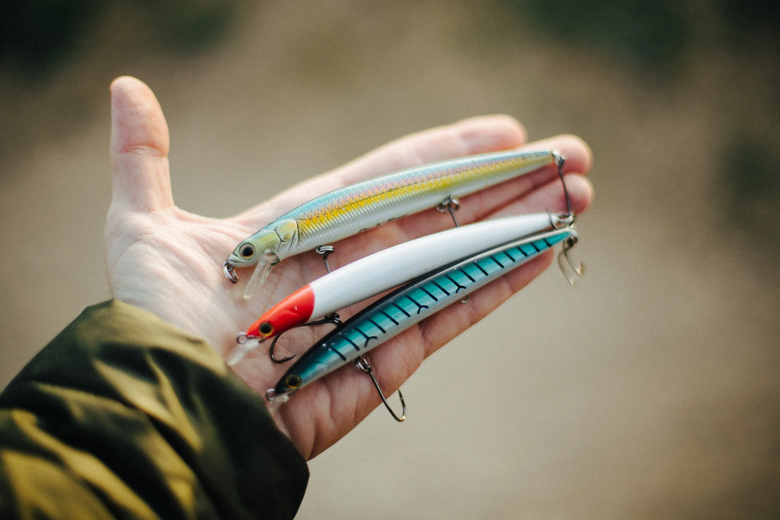 Three fishing lures held in a person's hand, with a blurred outdoor background.