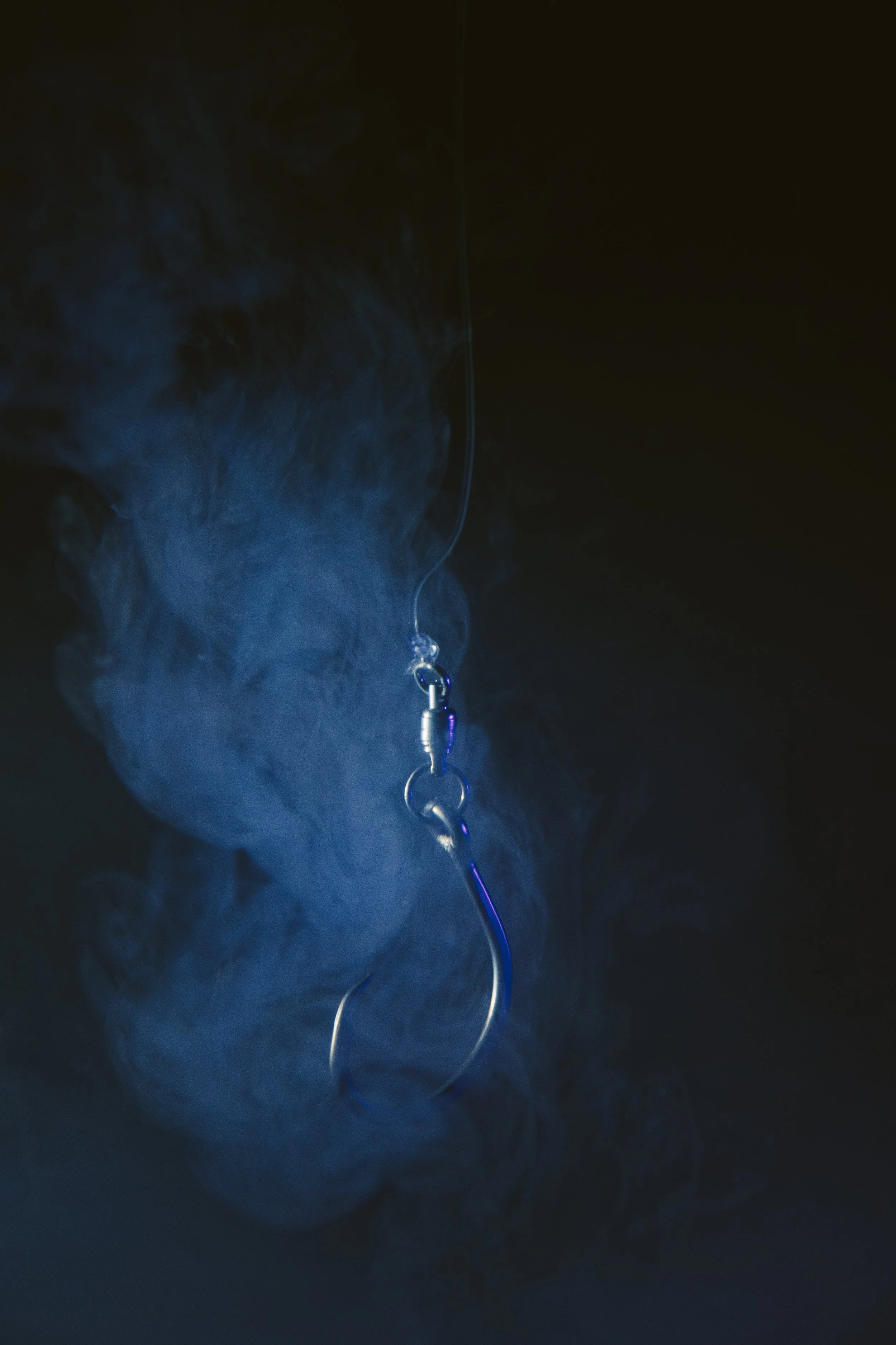 Close-up of a fishing hook hanging from a fishing line, surrounded by swirling smoke or fog in a dark environment.