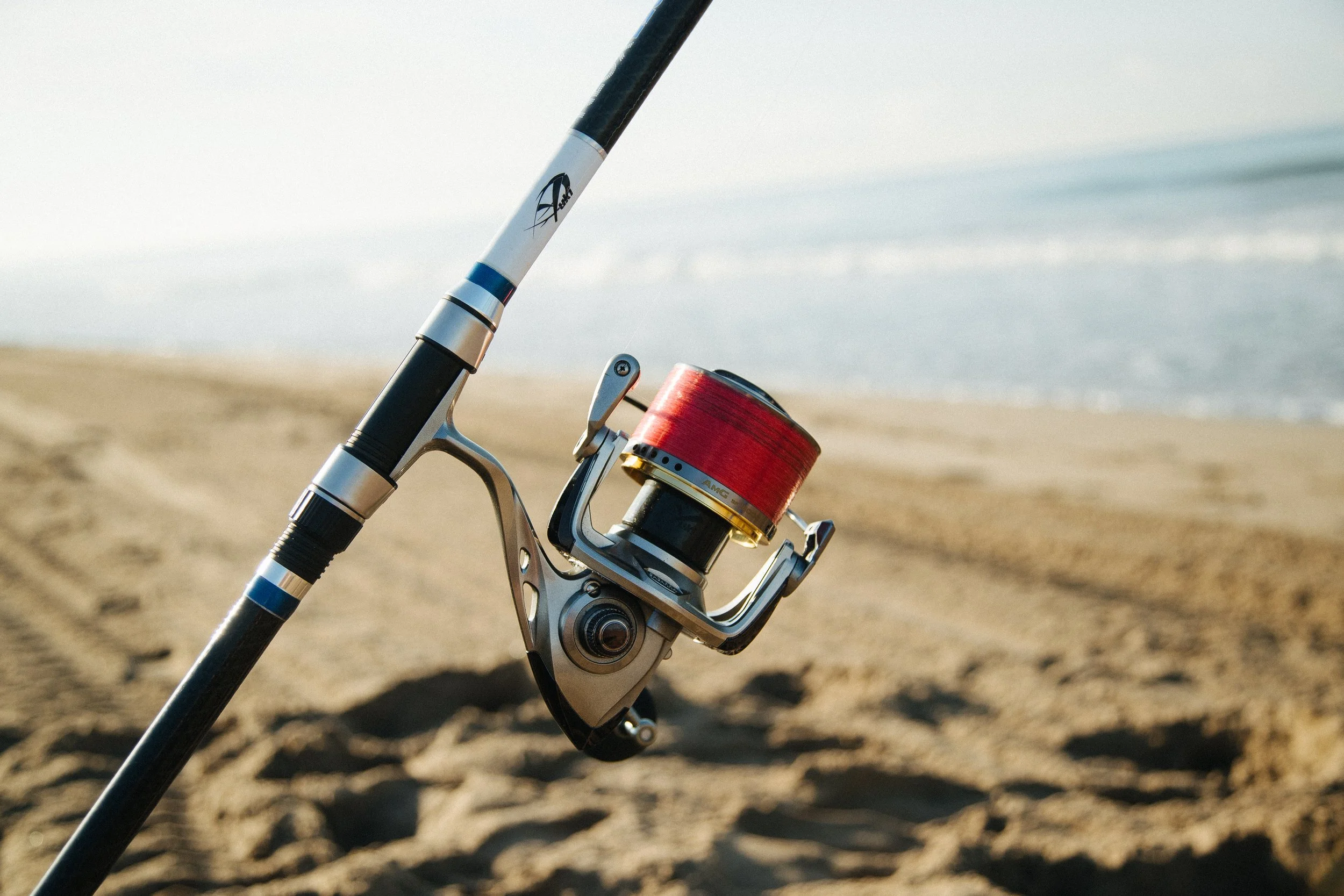 Fishing rod with reel stuck upright on sandy beach near water.