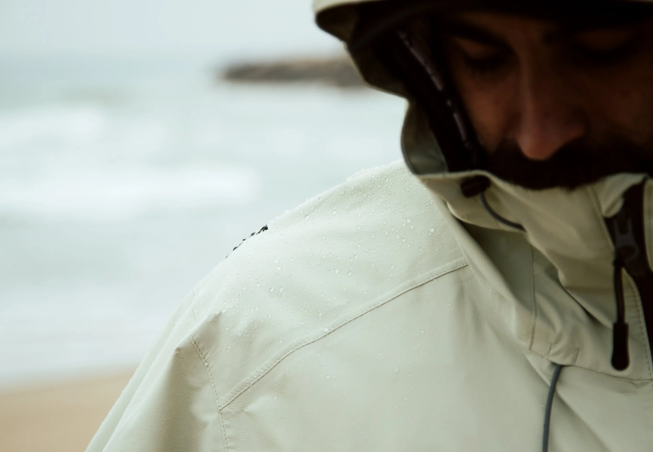 A person wearing a white rain jacket and black goggles, with water droplets on the jacket, near a beach with the ocean in the background.