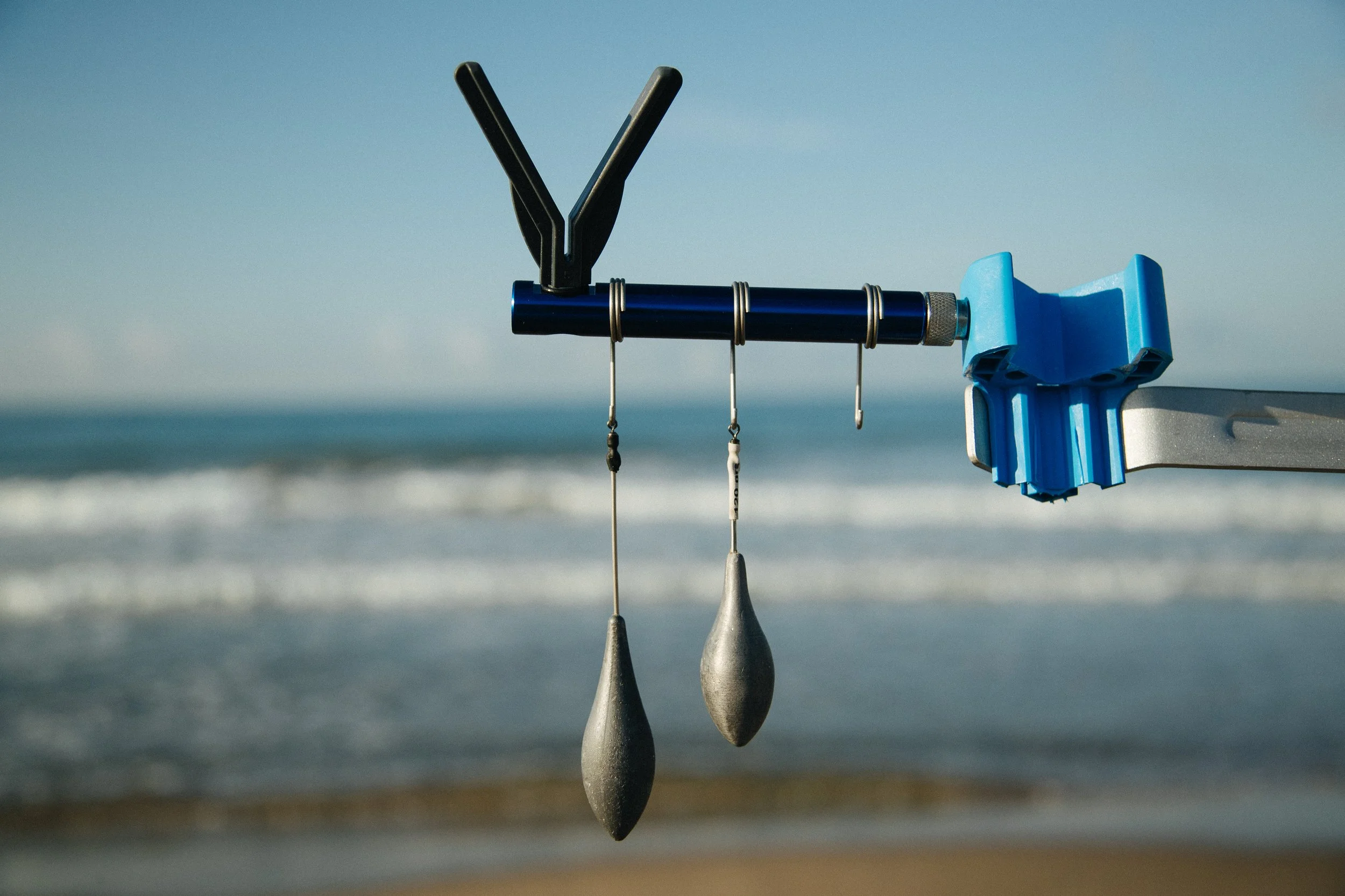 A metal rod with fishing weights hanging from it, mounted on a bracket against a beach background with ocean waves and a clear sky.