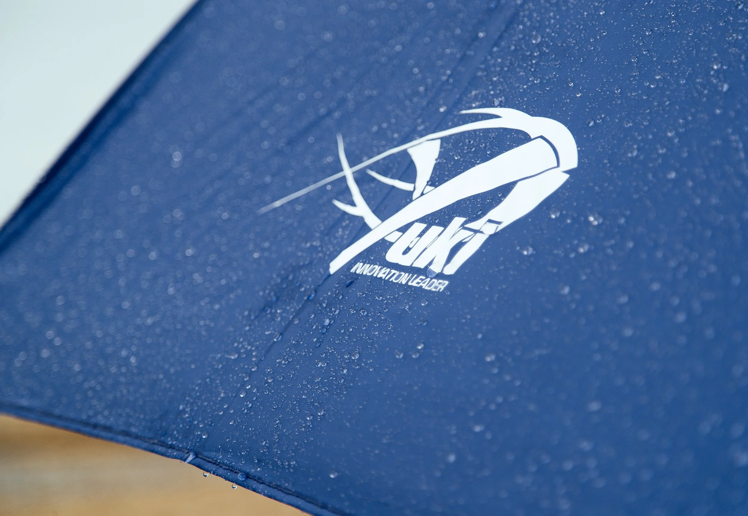 Close-up of a blue umbrella with water droplets on its surface and a white logo featuring a stylized shuttlecock and the text 'YONT' and 'INNOVATION LEADER'.