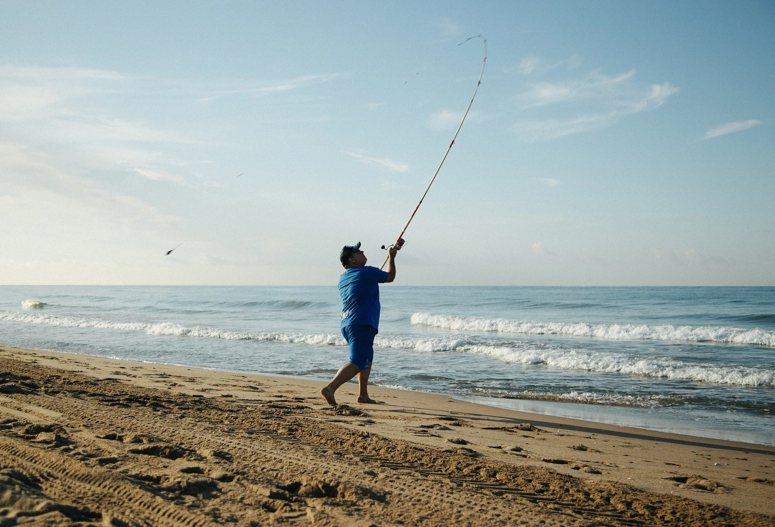 Man in blue shirt and shorts fishing on sandy beach with ocean waves and blue sky in the background.
