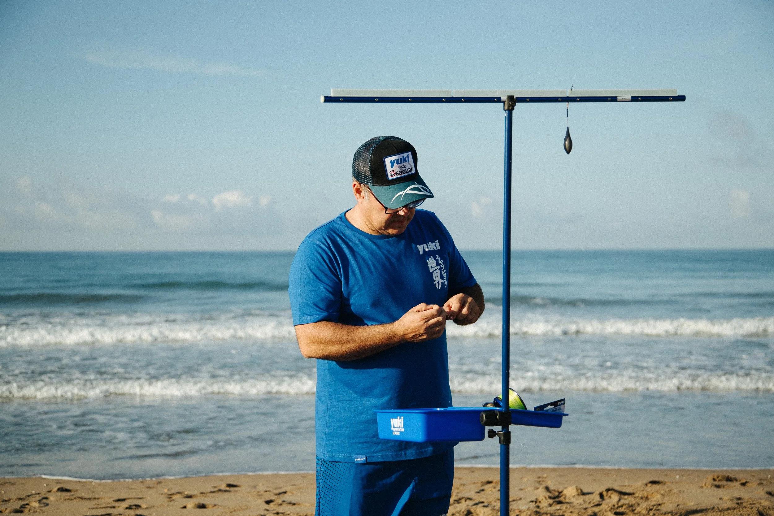 A man in a blue shirt and cap preparing fishing gear on a sandy beach with the ocean in the background.