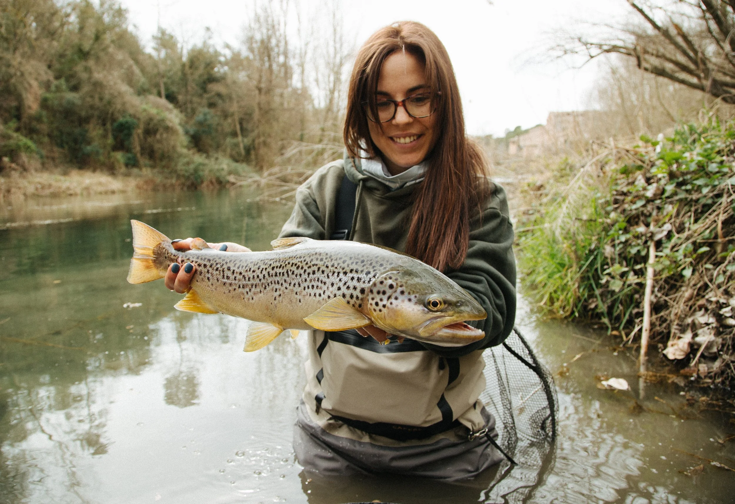 A woman in outdoor fishing gear holding a large trout fish she caught in a river.