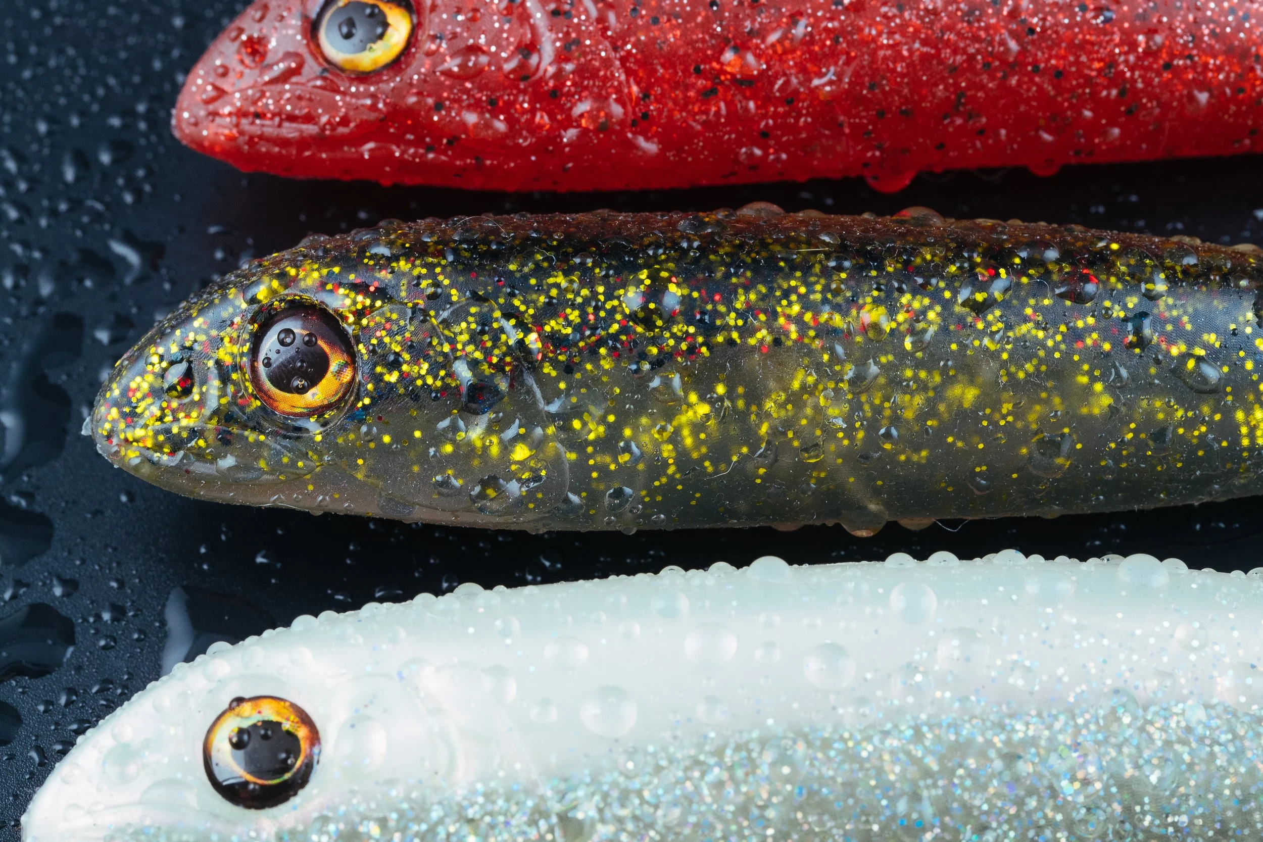 Close-up of three fishing lures shaped like fish, with water droplets on their surfaces. The top lure is red, the middle is black with yellow speckles, and the bottom is white with iridescent glitter.