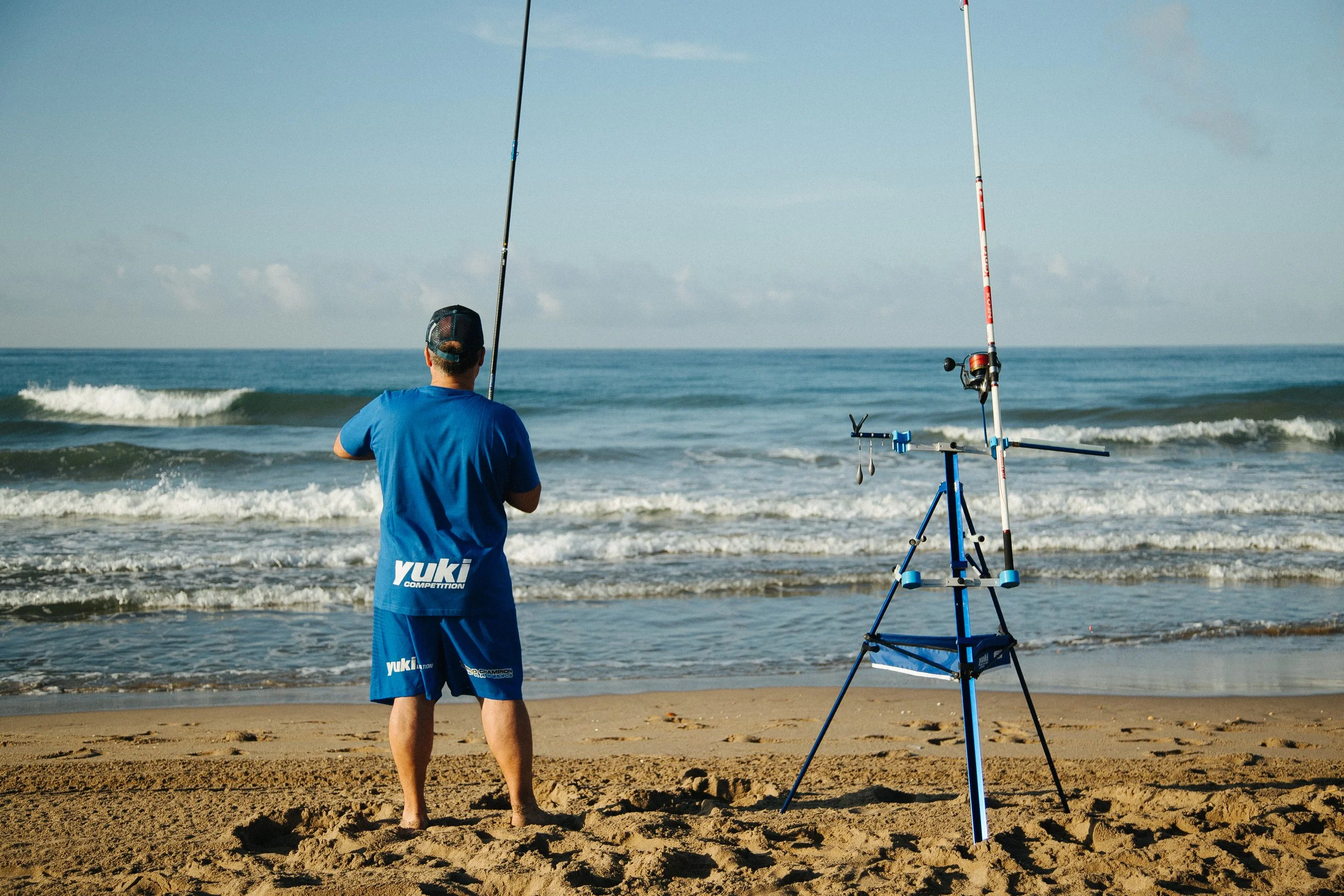 Person fishing on the beach with two fishing rods and a rod holder, facing the ocean with waves and a partly cloudy sky.