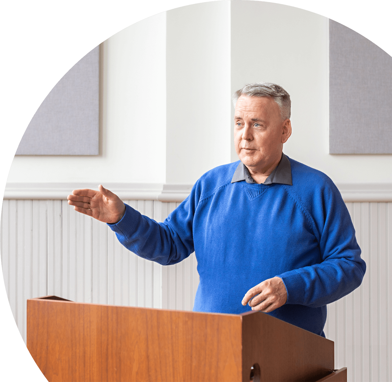 A middle-aged man with gray hair wearing a blue sweater and gray collared shirt standing at a wooden podium, speaking or presenting in a room with white wainscoting and gray acoustic panels on the wall.