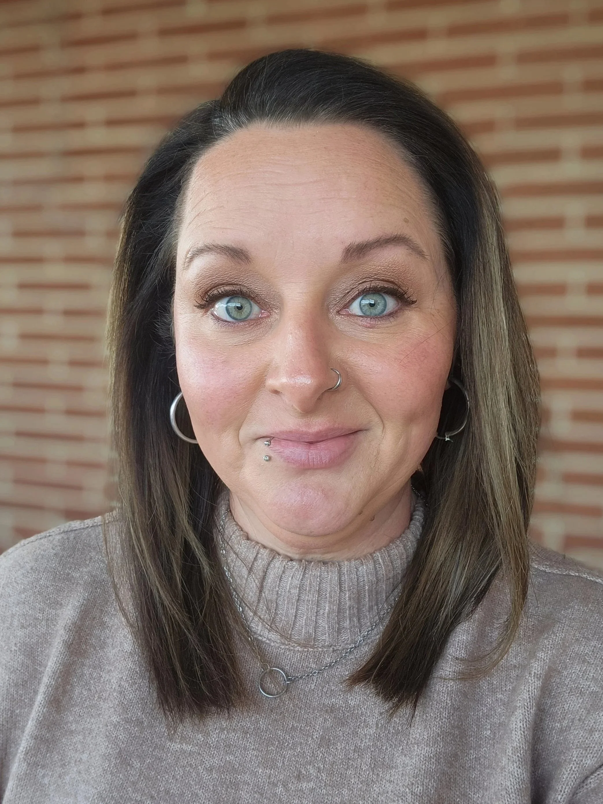 Close-up of a woman with blue eyes, dark brown hair with lighter highlights, wearing a beige sweater, silver hoop earrings, with lip and nose facial piercings, standing in front of a brick wall.