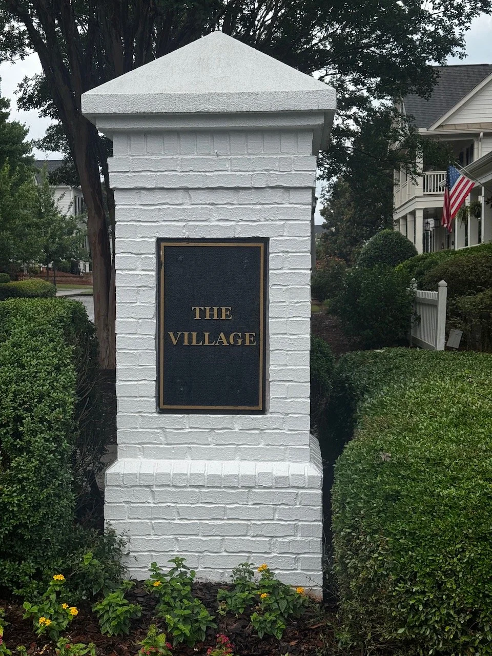 A brick building with white columns and a green metal roof, surrounded by neatly trimmed bushes and trees, under a partly cloudy sky with three flagpoles displaying American flags.