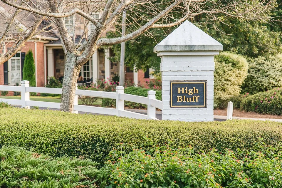A white brick post with a black and gold sign reading 'High Bluff' in front of a suburban house with a front yard, trees, and bushes.