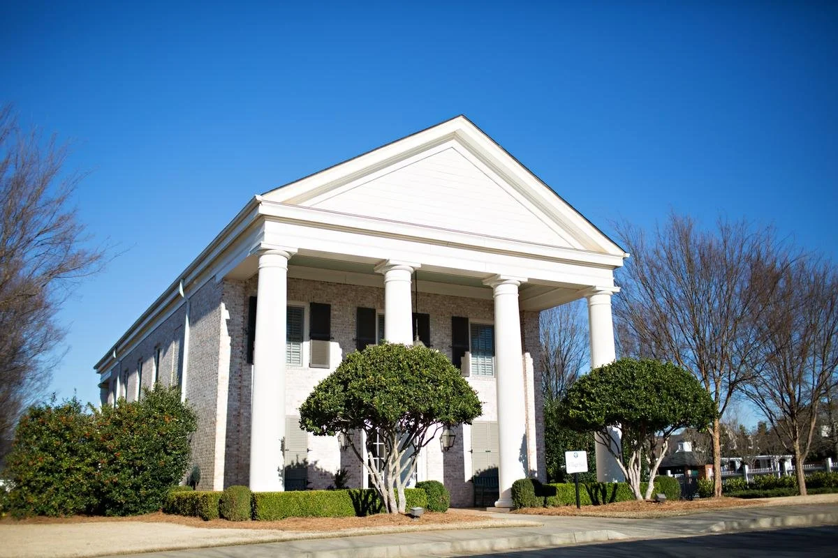 Georgia Club  large, two-story white brick building with a front porch supported by four white columns, surrounded by manicured bushes and trees under a clear blue sky.