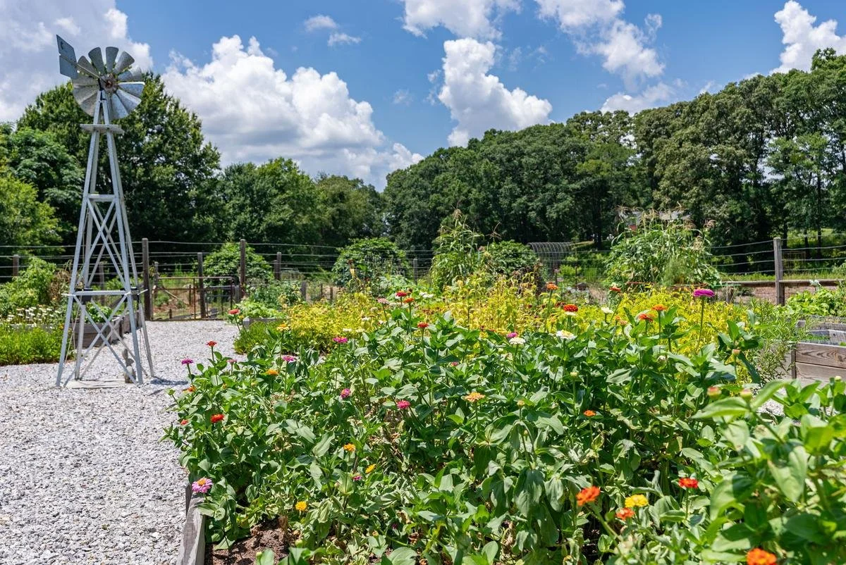 A garden with a metal windmill, colorful flowers, and lush trees in the background under a partly cloudy sky.