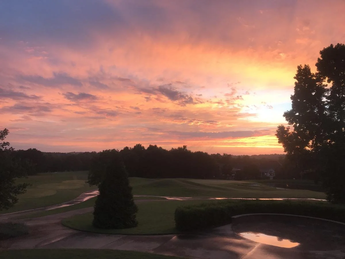 Sunset with pink, orange, and purple clouds over a golf course, trees, and water reflections.