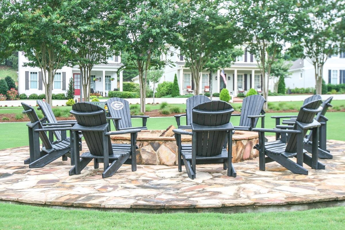 A circular stone fire pit with eight black wooden Adirondack chairs arranged around it, set on a stone patio in a landscaped yard with trees, grass, and houses in the background.