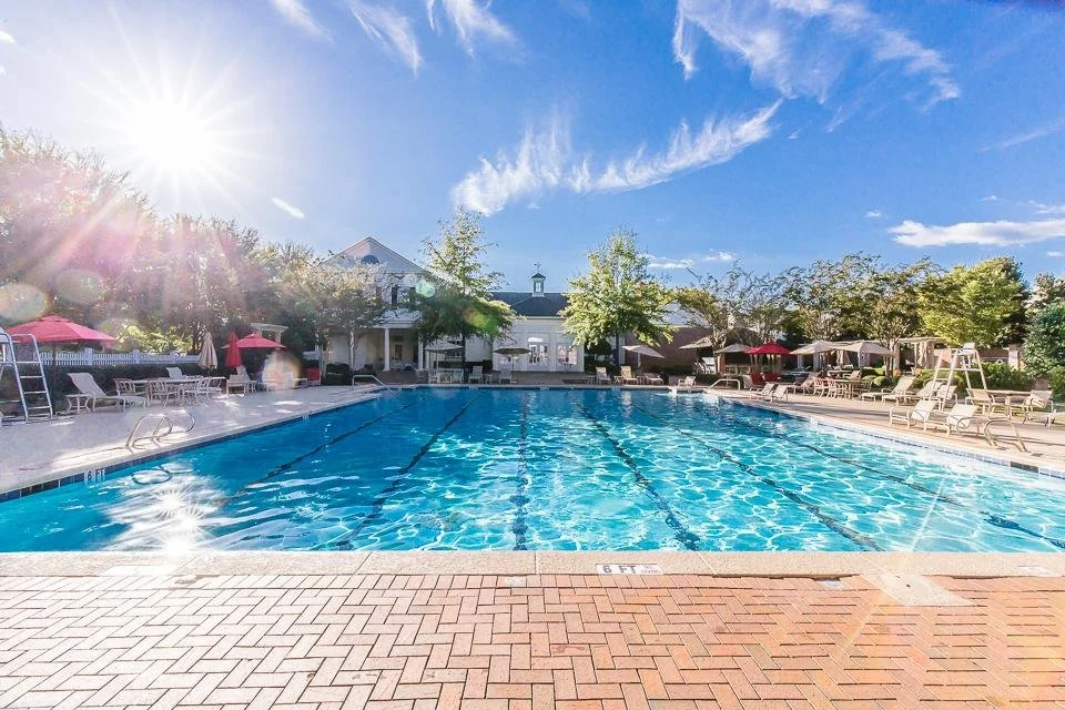 Outdoor swimming pool surrounded by lounge chairs, umbrellas, and trees under a sunny sky.