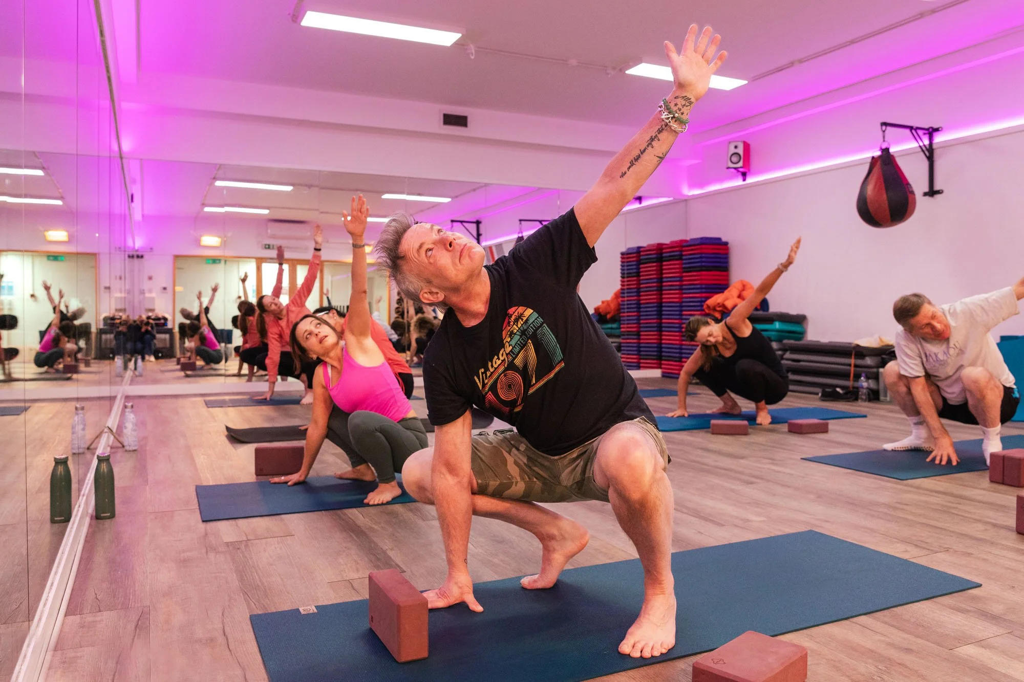 An indoor yoga class with participants on mats performing yoga poses, mirrored walls, and a gym-like setting.