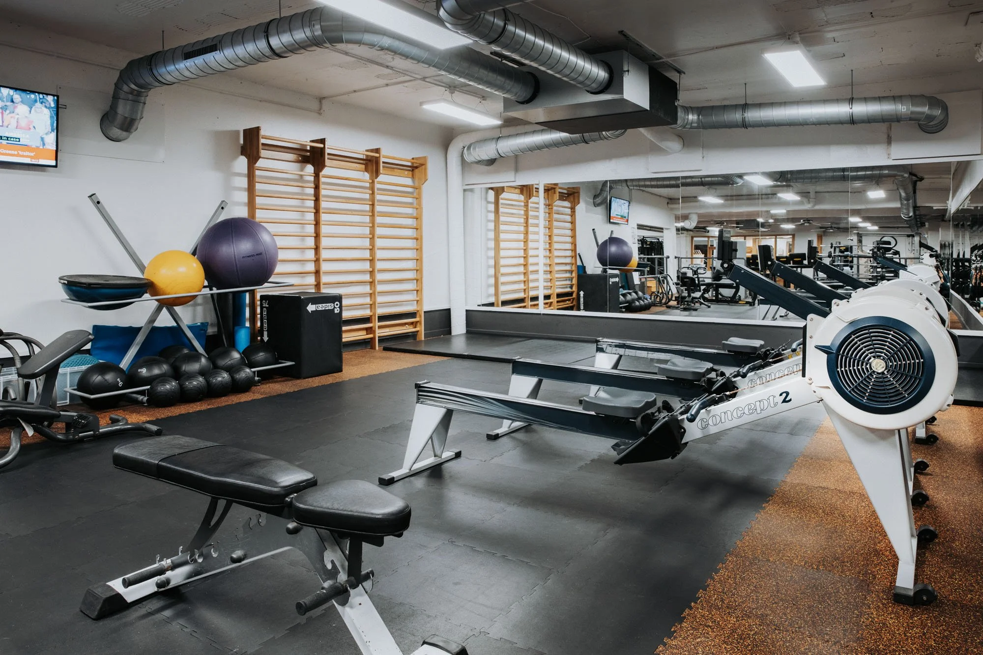 Fitness studio with rowing machines, weight rack, medicine balls, and climbing wall with wooden bars. Large mirror on wall reflecting equipment.
