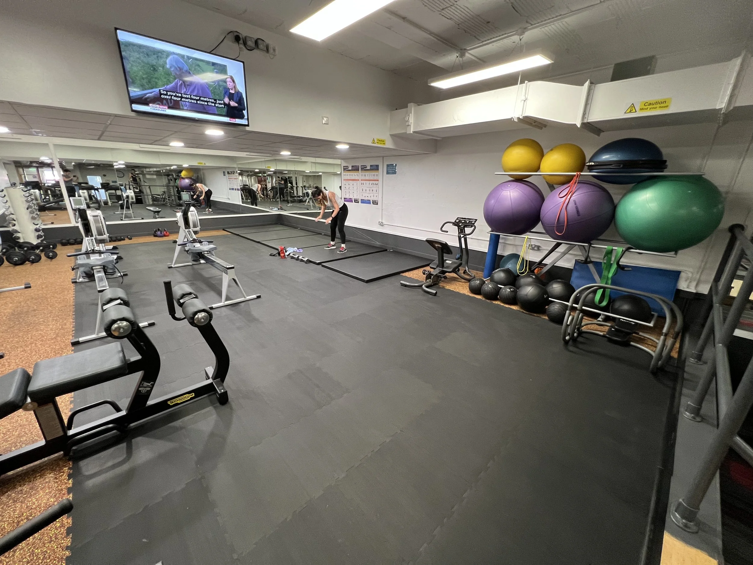 A gym with exercise equipment including rowing machines, a station with resistance bands and battle ropes, and shelves holding large exercise balls and smaller workout balls. A woman is preparing to exercise, and a large mirror covers one wall. A television is mounted on the wall above.