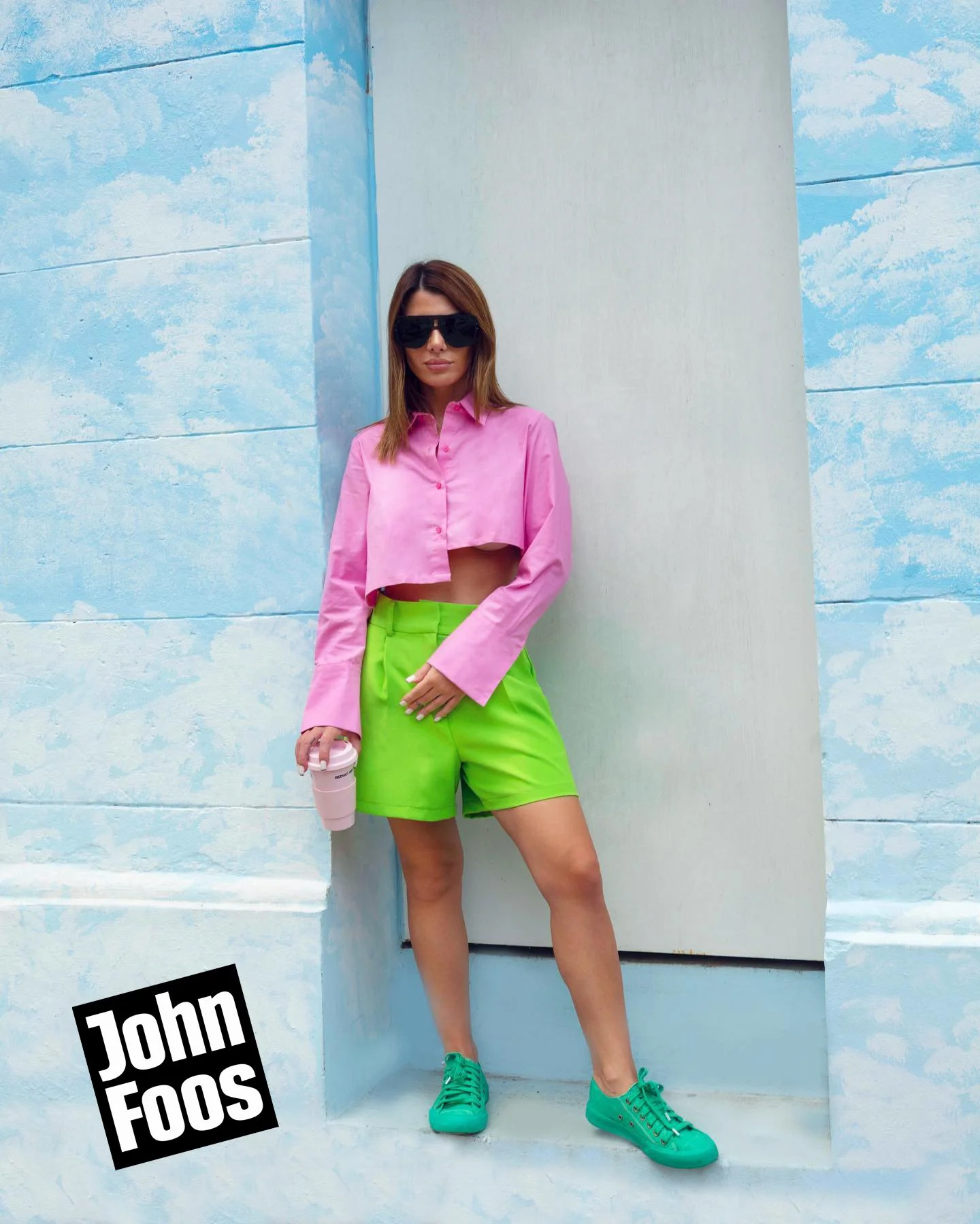 Mujer con gafas oscuras, camisón rosa corto, pantalones cortos verdes y zapatos deportivos verdes, sosteniendo un vaso rosa, de pie frente a una pared de colores pastel en un entorno urbano