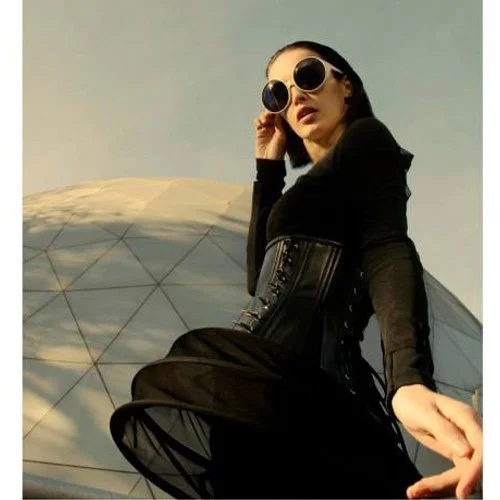 Una mujer con gafas grandes y de color oscuro, con cabello oscuro y peinado hacia atrás, viste ropa negra y está posando en frente de una estructura metálica moderna y una cancha en un día soleado