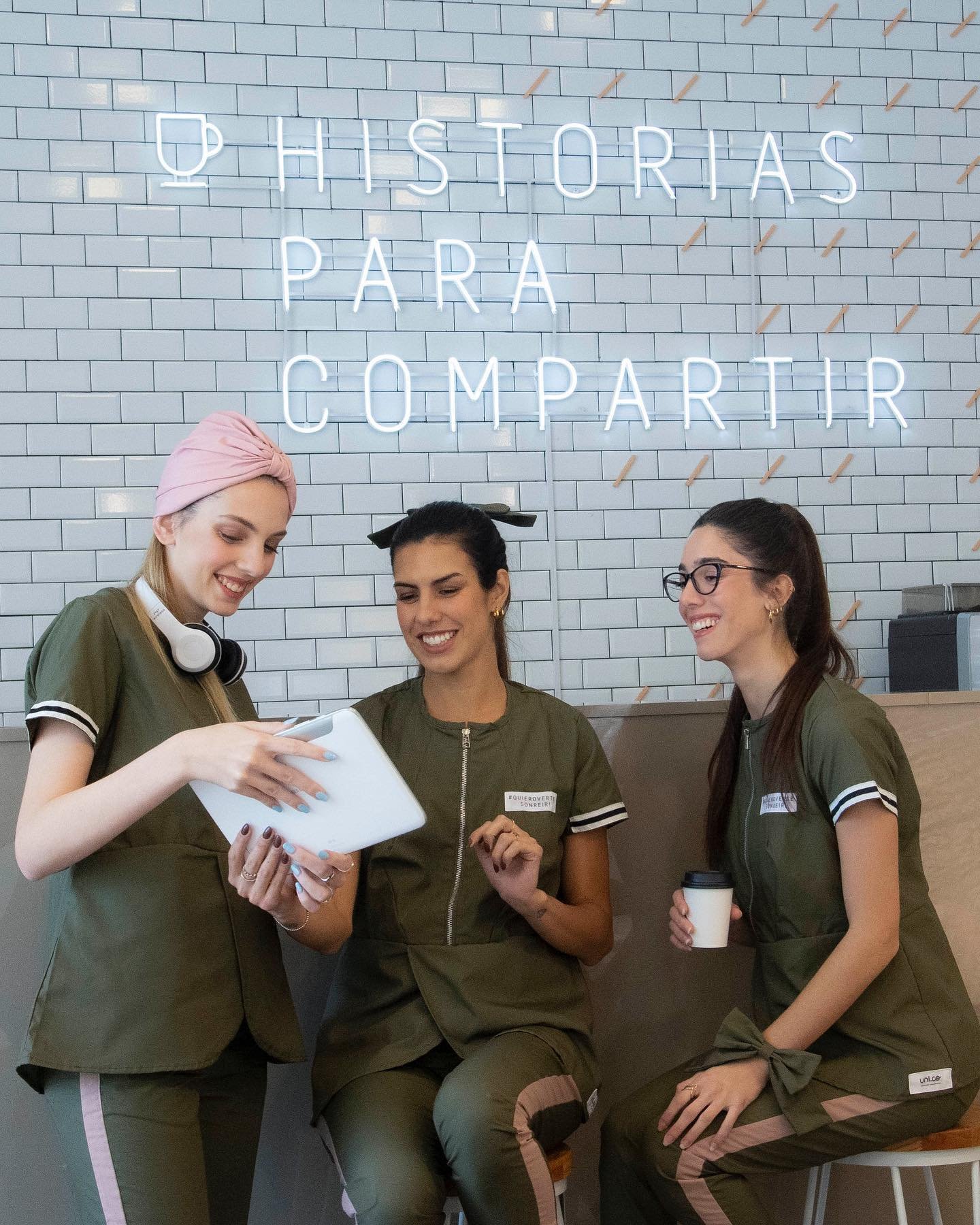 Tres mujeres sonriendo y mirando una tableta en un lugar con pared de azulejos y un letrero de neón que dice "Historias para Compartir".