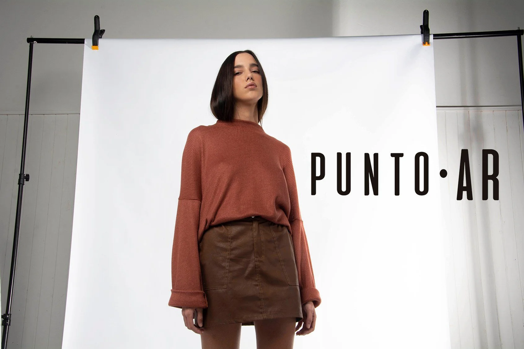 Mujer vistiendo un conjunto en tonos marrón en un estudio fotográfico, con un cartel que dice 'PUNTO AR'.