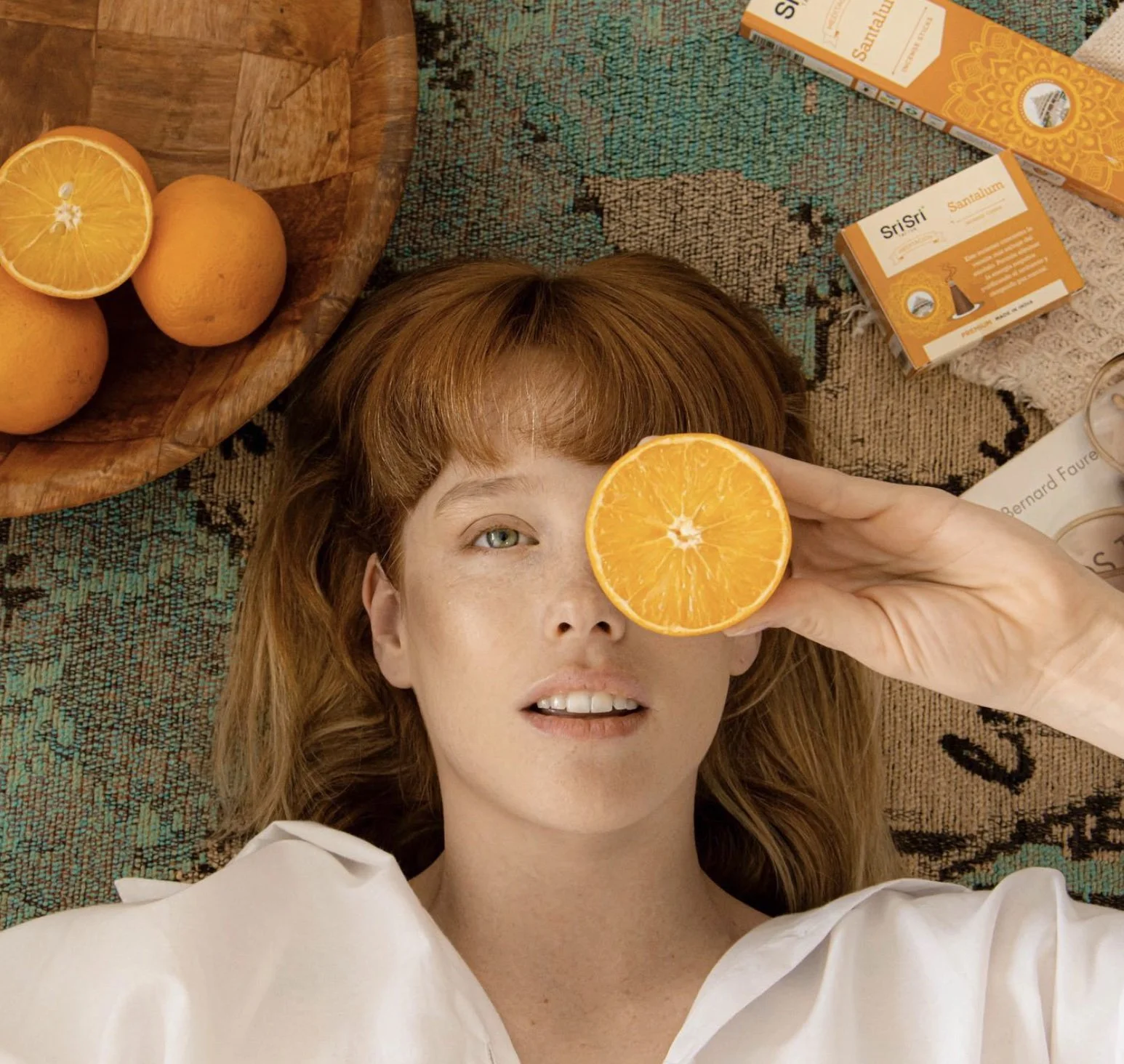 Mujer pelirroja con ojos claros acostada en un tapete, sosteniendo una rodaja de naranja frente a un ojo, rodeada de naranjas, cajas de tés y otros objetos en la superficie.