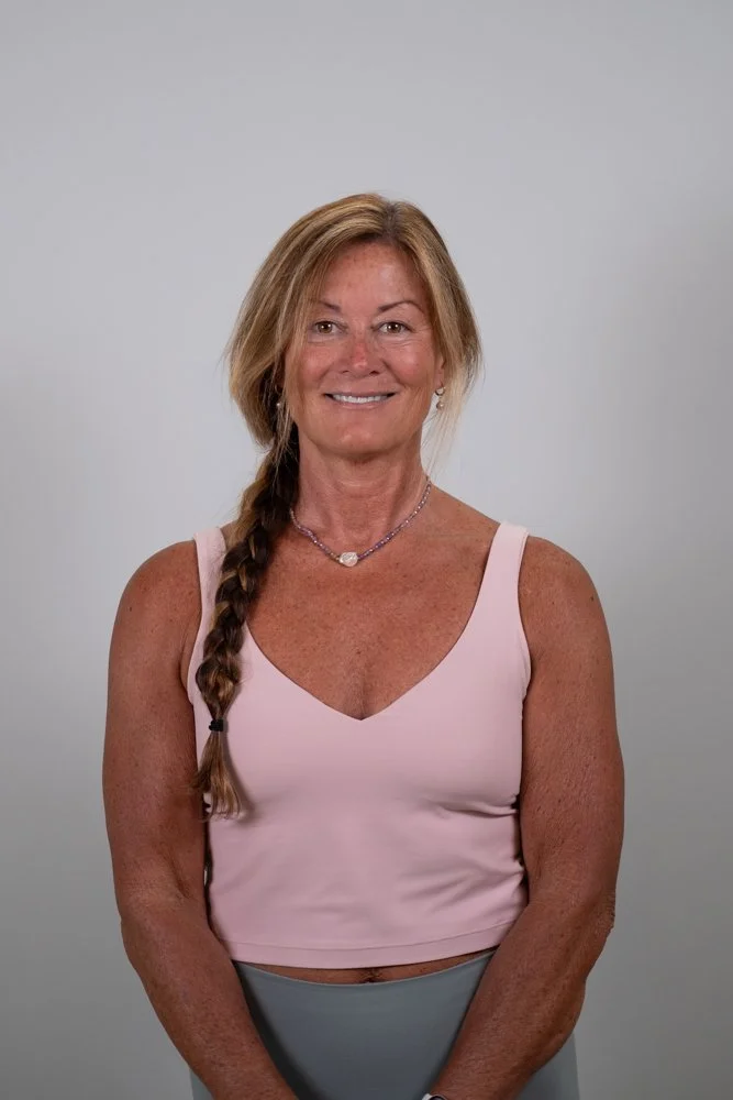 A smiling woman with long brown hair wearing a white tank top and black patterned leggings stands in front of a turquoise wall with white floral and geometric designs.