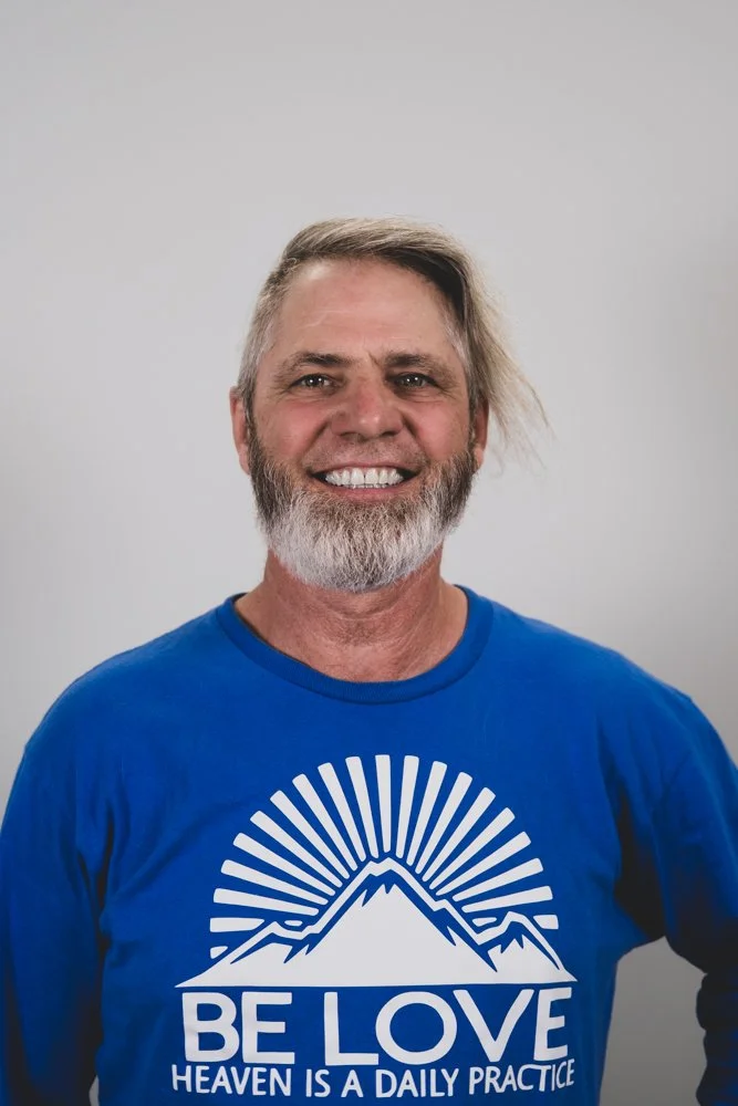 A smiling middle-aged man with gray hair and beard, wearing a dark t-shirt and a beaded bracelet, standing with arms crossed against a plain light background.