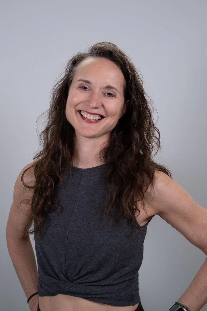 A woman with short, curly brown hair smiling at the camera, wearing a dark gray tank top and a necklace, standing against a plain white background.