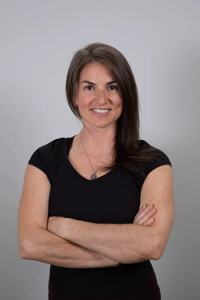 Smiling woman with long brown hair in a braid, wearing a blue sleeveless athletic shirt and a beaded necklace, standing against a plain light gray background.