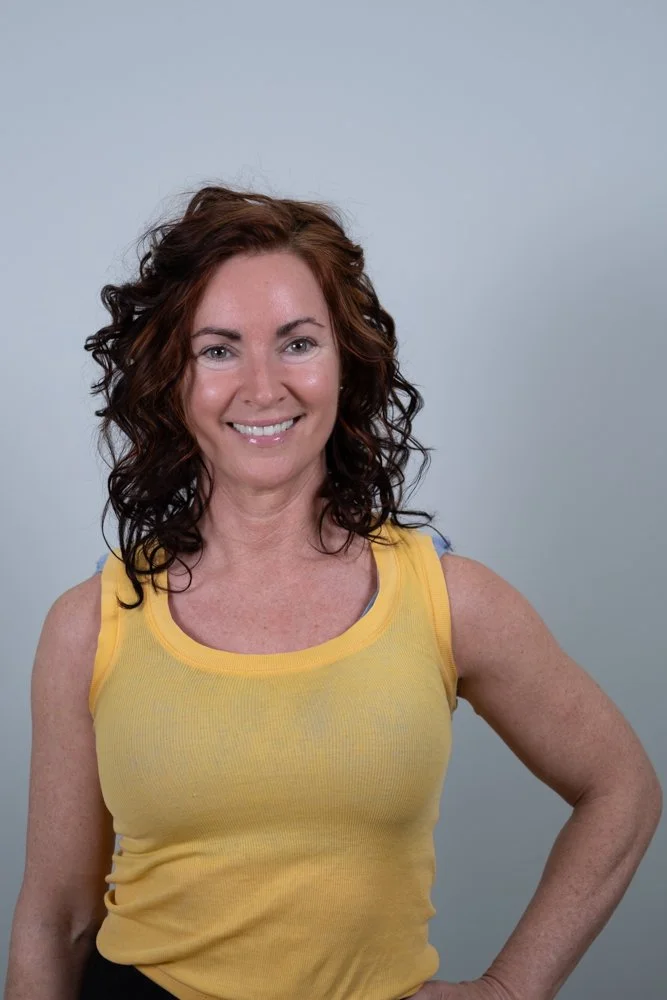 A woman with curly brown hair wearing a sleeveless blue tank top and red pants, standing in front of a large abstract logo with mountains and geometric shapes on a light blue background.