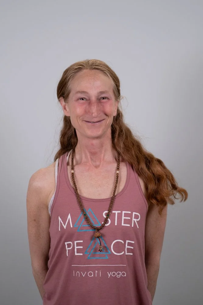 A woman with long, wavy red hair smiling at the camera, wearing a purple sleeveless top and a long beaded necklace, standing against a plain light-colored background.