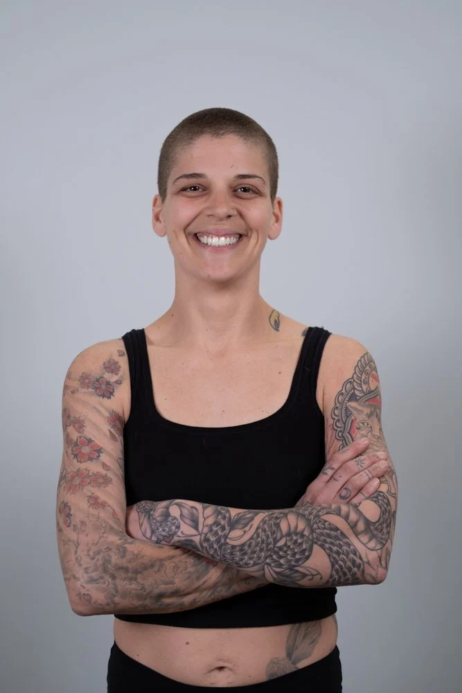 Young woman with a short haircut smiling and wearing a black tank top, showing tattoos on her left arm, against a plain light background.