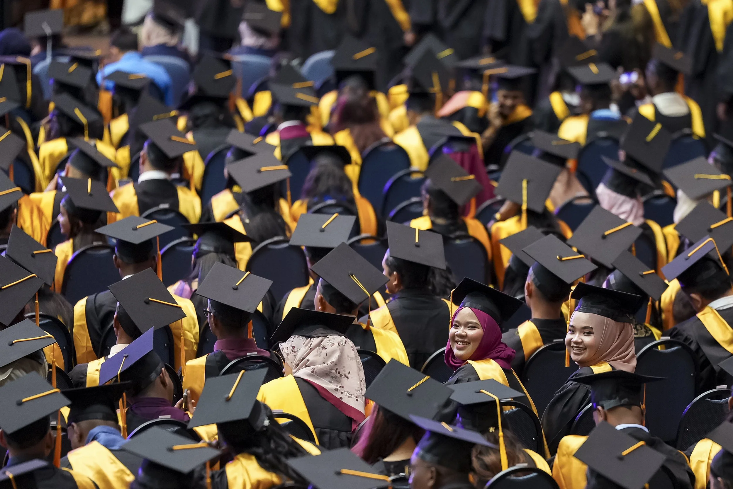 Large group of graduates wearing black caps and gowns with yellow stoles and mortarboards, seated during a graduation ceremony. Some students are smiling, and a few women wearing colorful hijabs are visible in the crowd.